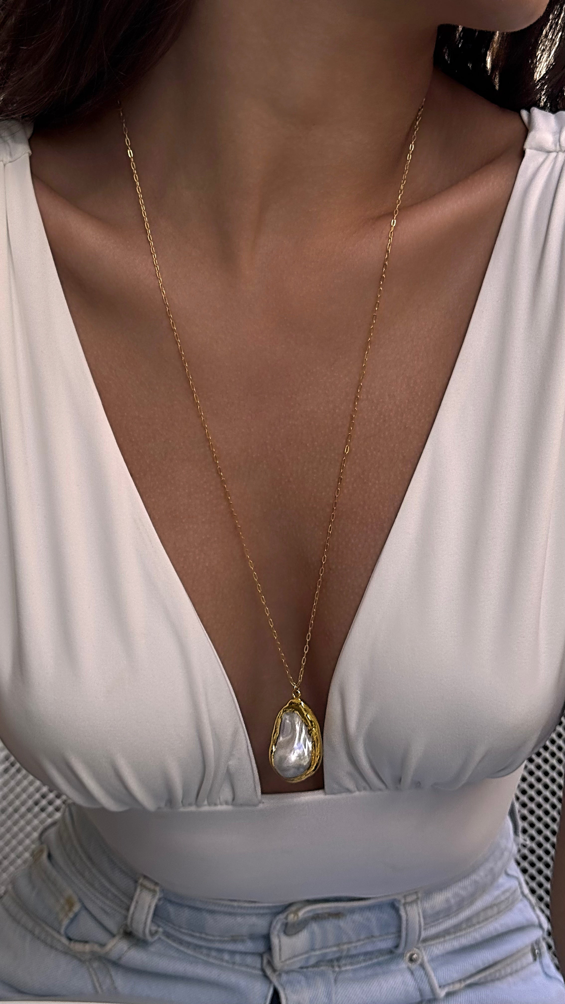Woman wearing a gold-plated necklace with a pearl pendant, dressed in a white top.