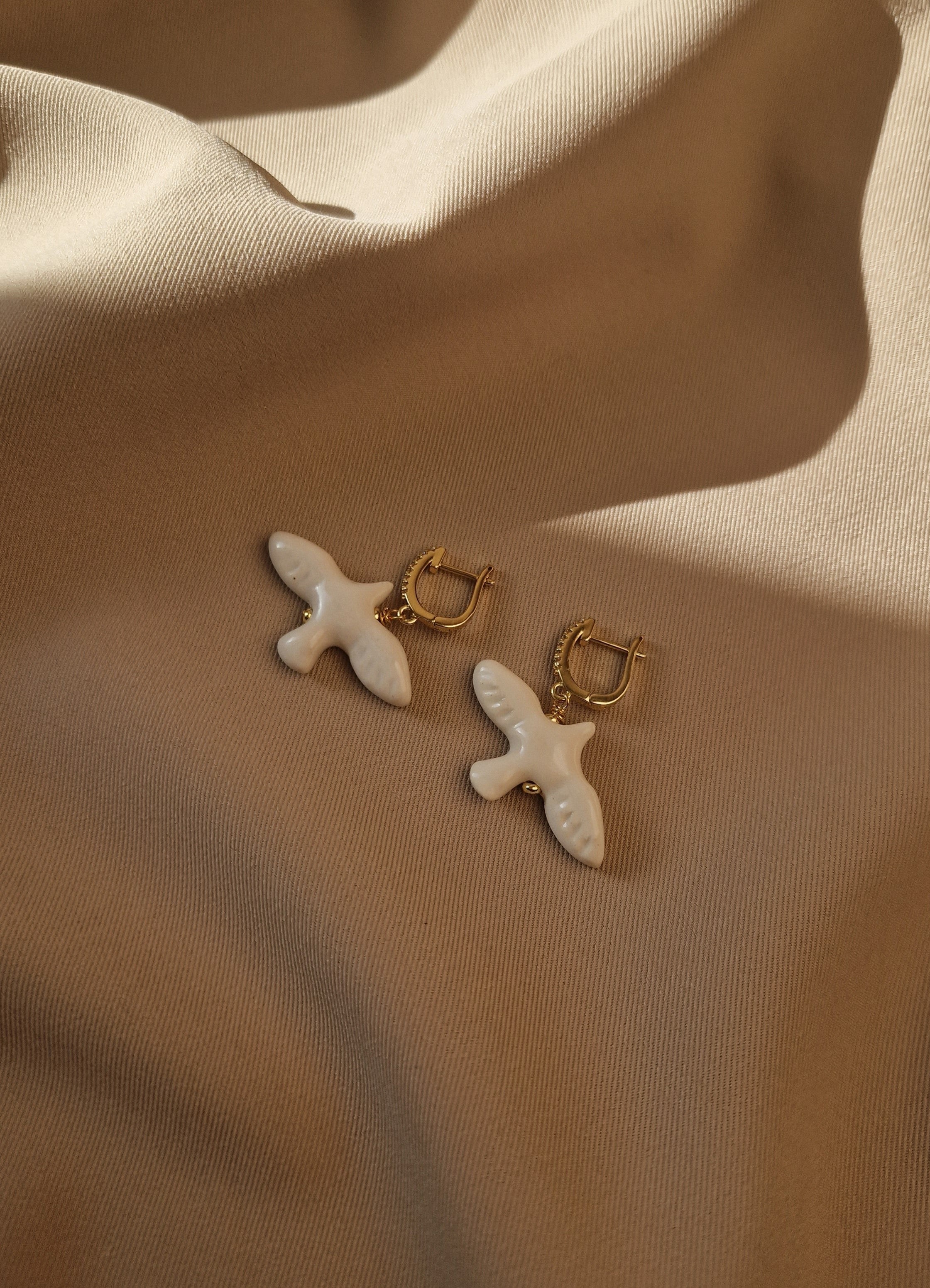 Pair of earrings with gold and white bird designs on a beige fabric background