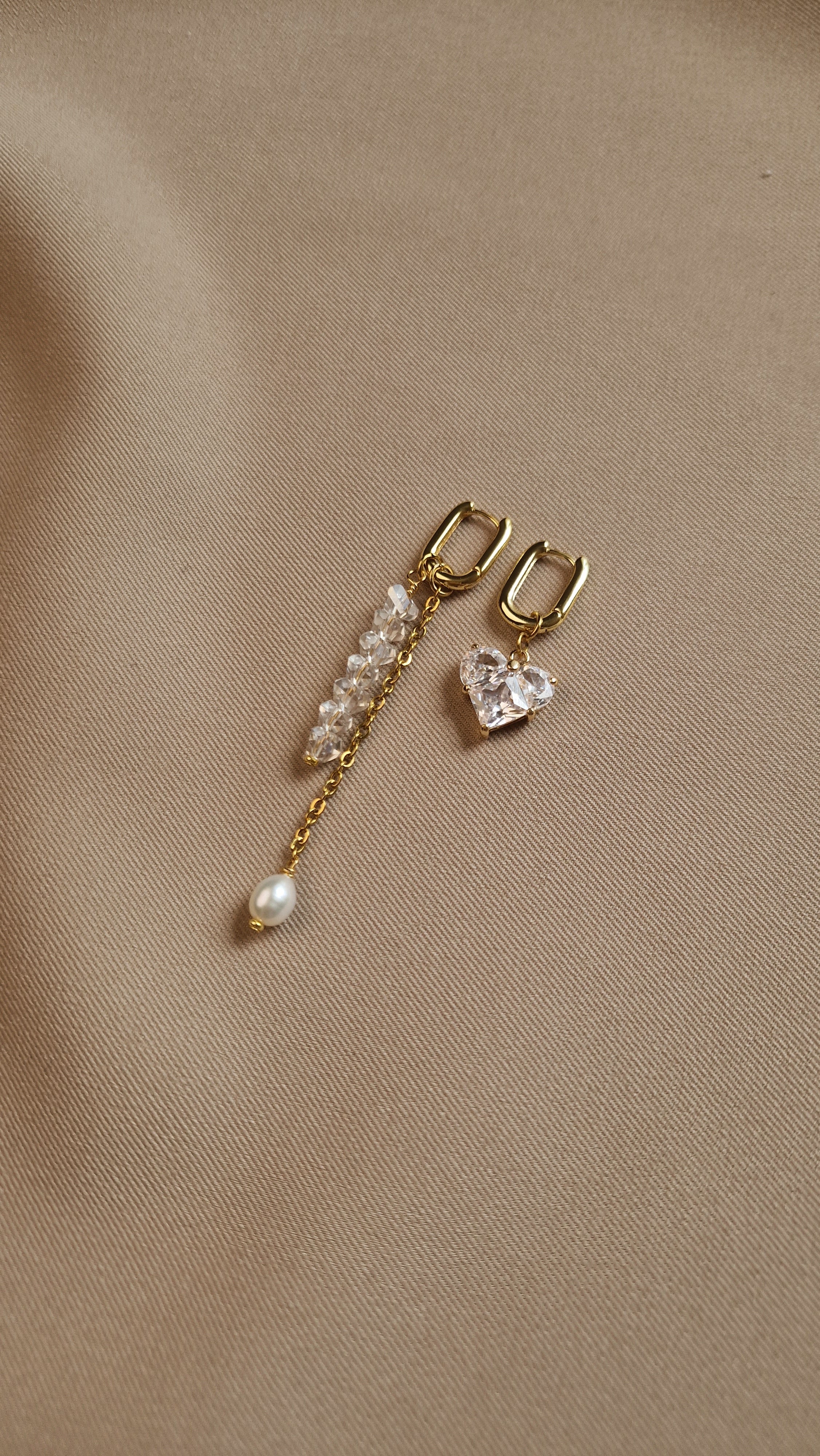 Gold-plated earrings with heart and pearl details on a beige fabric background