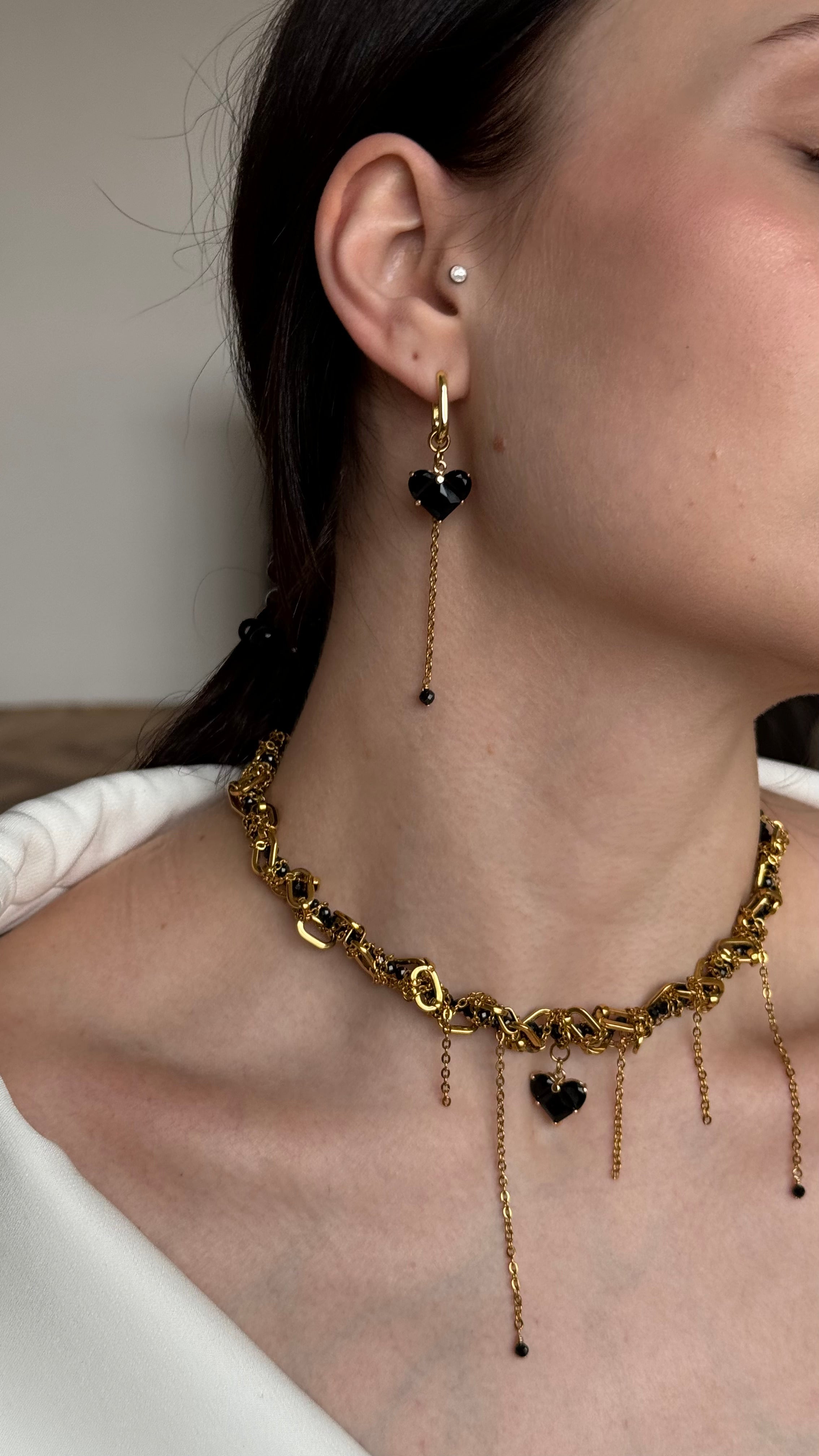 Gold-plated necklace and earings  with black heart pendants worn by a person in a white outfit.