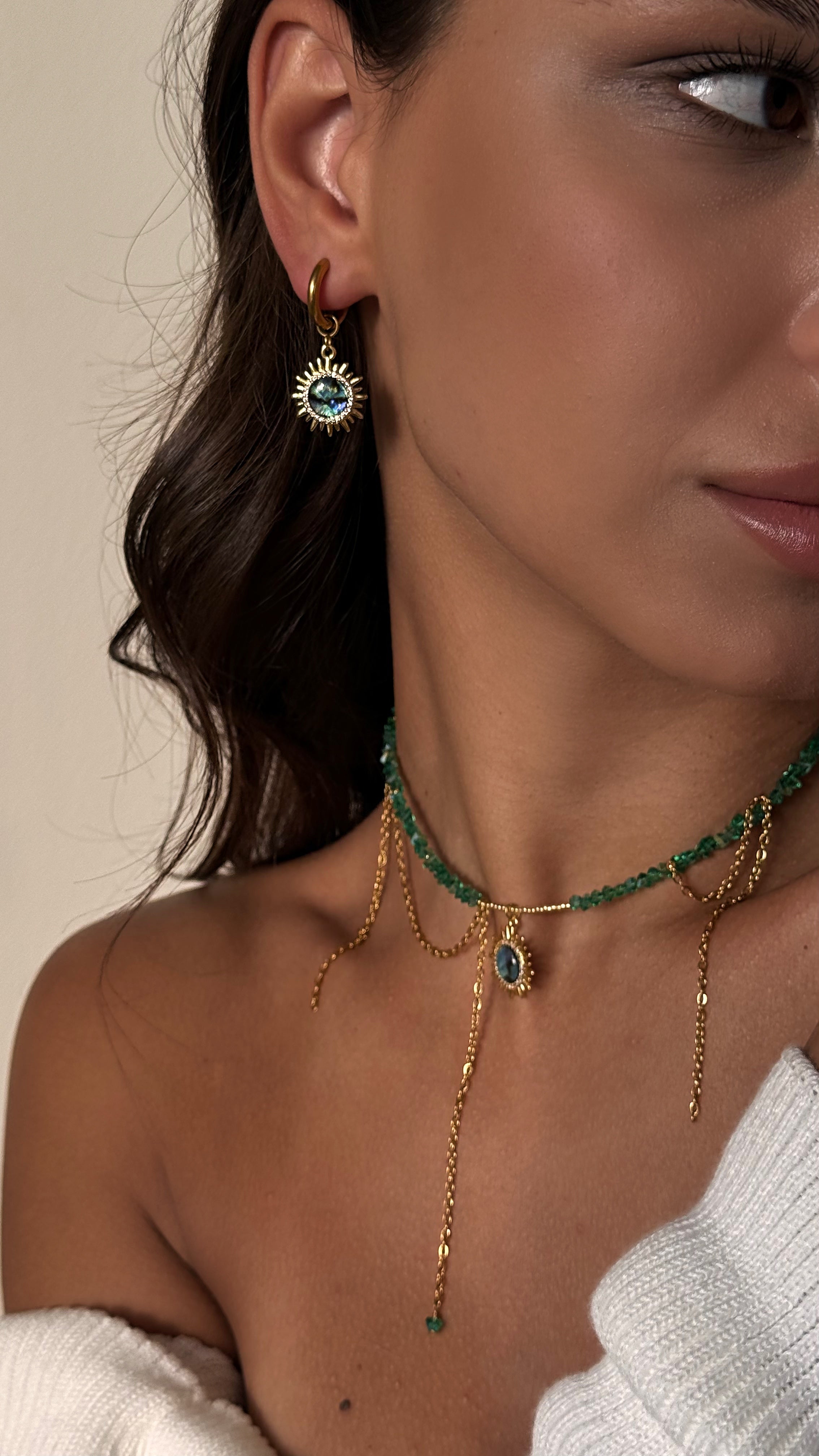 Close-up of a woman wearing gold-plated earrings and a green necklace against a beige background