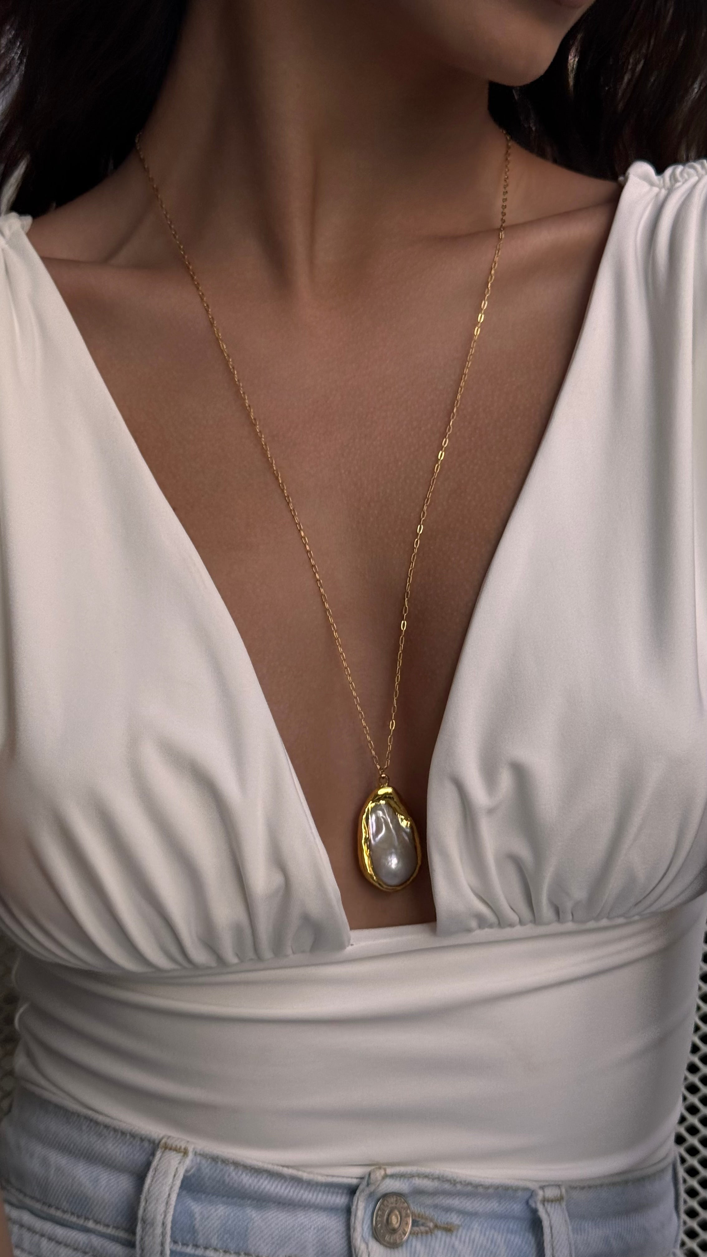 Woman wearing a gold-plated necklace with a pearl pendant, dressed in a white top.