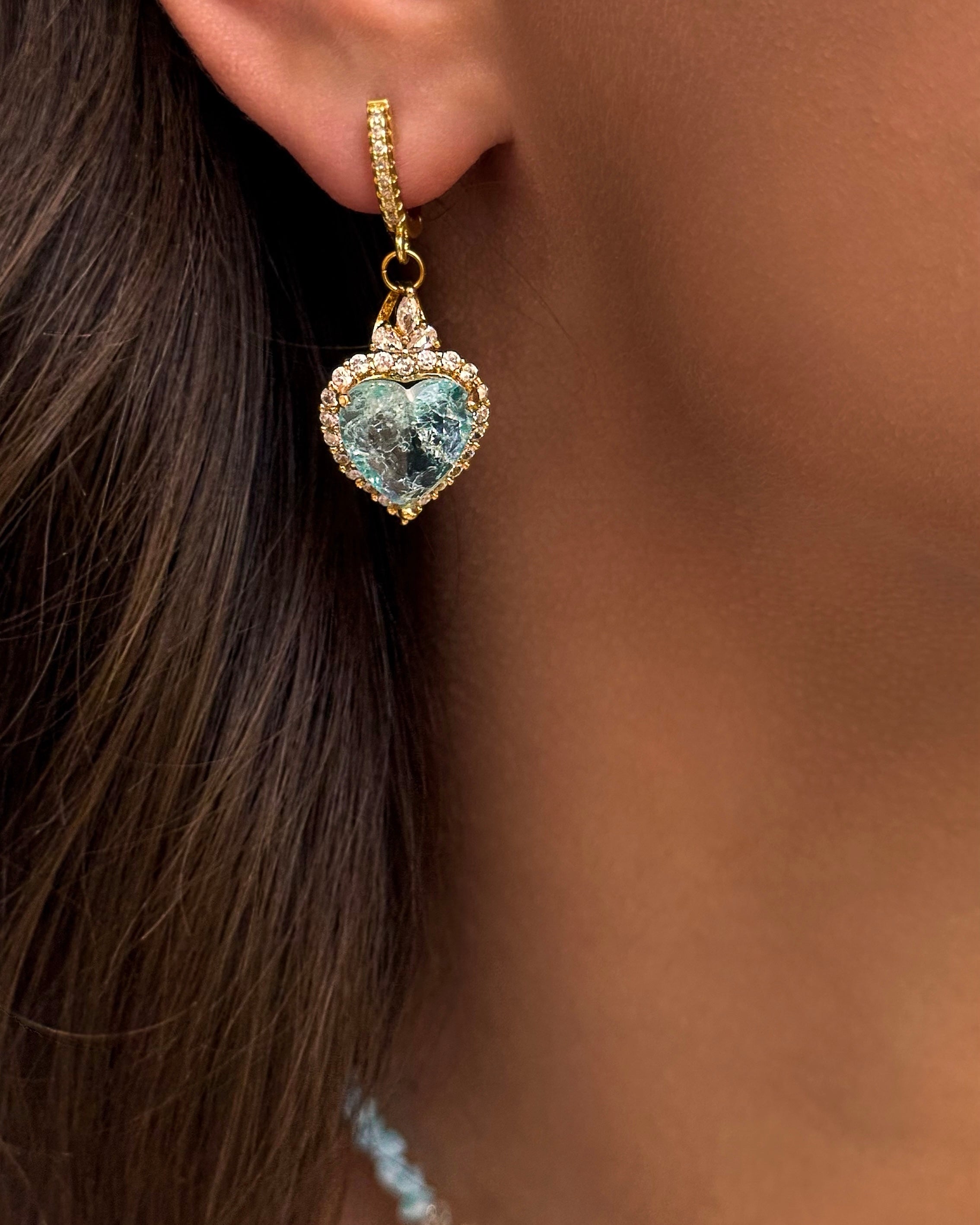 Gold-plated heart-shaped earring with a blue gemstone worn by a person.
