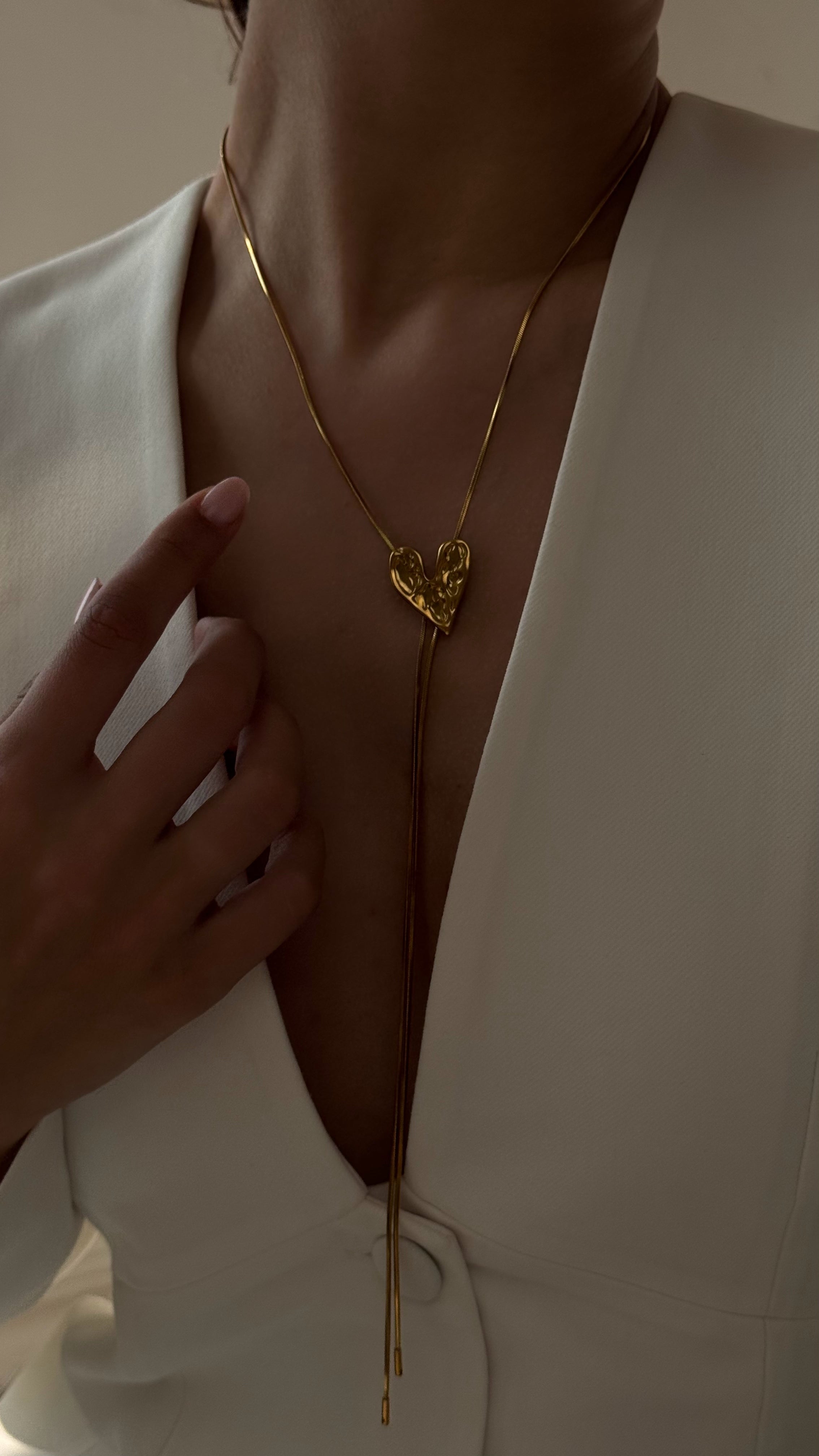Gold-toned jewelry set with a long heart pendant necklace, matching heart earrings, and an adjustable heart ring, shown on a model styled in a chic, art deco-inspired look.